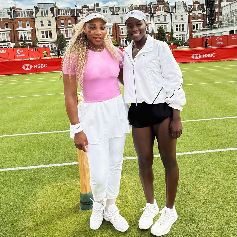 Tennis legend Serena Williams and player Victoria Mboko pose together for a photo on an outdoor grass tennis court. Serena, on the left, smiles while wearing a pink short-sleeved top, a white tennis skirt over white athletic pants, white Nike shoes, and a white visor. Victoria Mboko, on the right, smiles alongside her, dressed in a white Wilson zip-up jacket, black athletic shorts, white tennis shoes, and a white visor. They are standing on the green grass court under a bright, partly cloudy sky, with red HSBC sponsor banners and row houses visible in the background.