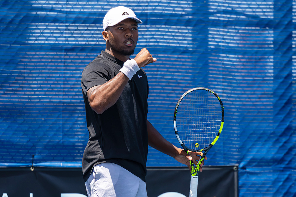 Tennis player Jean-Baptiste Badon celebrates a point with a fist pump during a match at the SoCal Pro Series in Lakewood. He is wearing a black Nike t-shirt, grey shorts, a white Adidas cap, and a white wristband on his right arm. He holds his yellow and black tennis racket in his left hand, looking forward with intense focus. The background features a textured blue windscreen on a bright, sunny day.