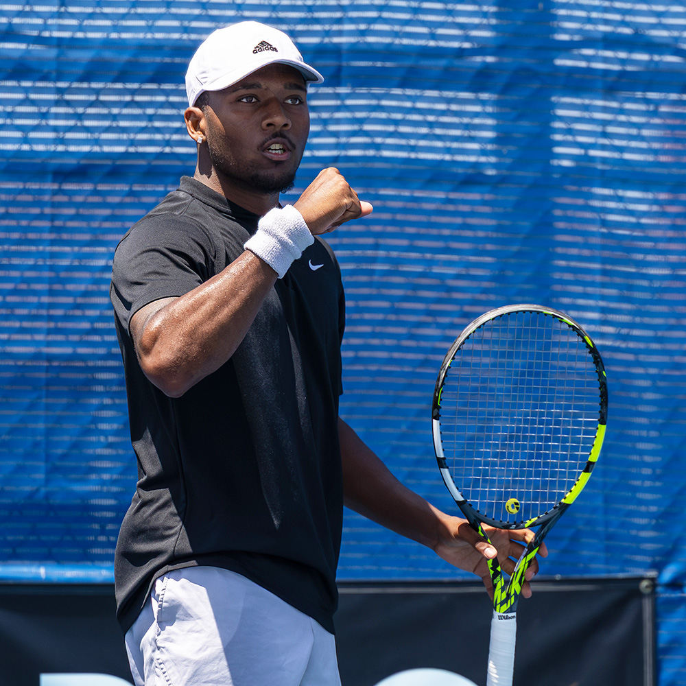 Tennis player Jean-Baptiste Badon celebrates a point with a fist pump during a match at the SoCal Pro Series in Lakewood. He is wearing a black Nike t-shirt, grey shorts, a white Adidas cap, and a white wristband on his right arm. He holds his yellow and black tennis racket in his left hand, looking forward with intense focus. The background features a textured blue windscreen on a bright, sunny day.