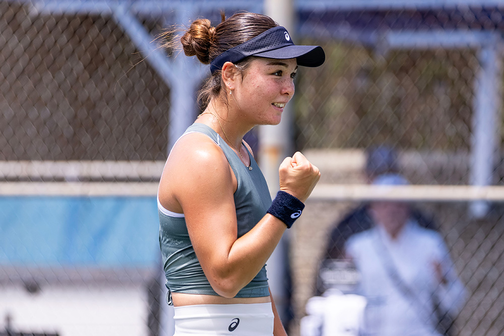 UCLA Bruins tennis player Mayu Crossley celebrates on court after defeating former Junior World No. 1 Kristina Penickova at the SoCal Pro Series in Lakewood. She is captured smiling in profile with her right fist pumped forward in celebration. She is wearing a grey Asics tank top, a white tennis skirt, a dark blue Asics visor, and a matching blue wristband. A chain-link fence on a sunny outdoor court frames the shot, with a blurred figure visible in the background.