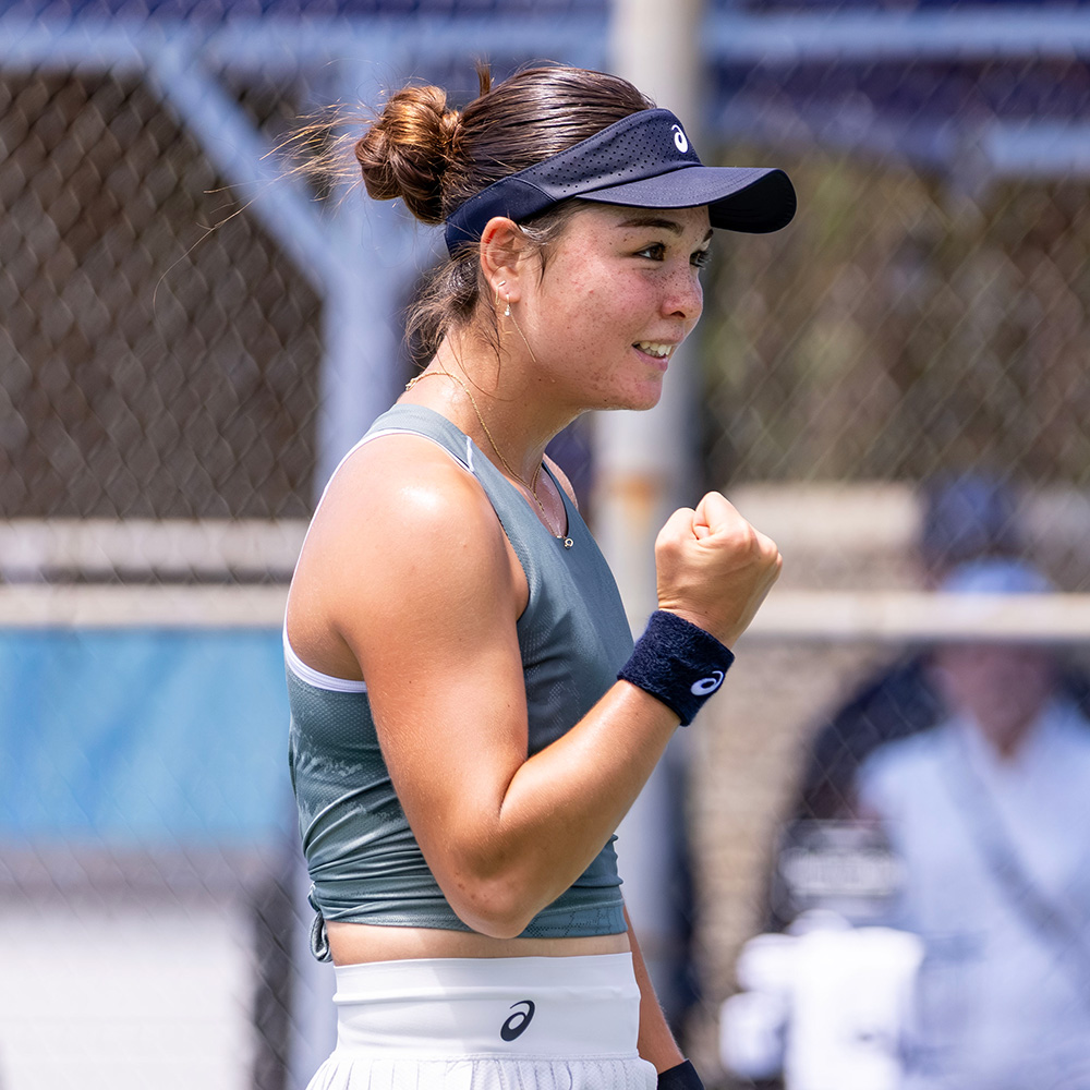 UCLA Bruins tennis player Mayu Crossley celebrates on court after defeating former Junior World No. 1 Kristina Penickova at the SoCal Pro Series in Lakewood. She is captured smiling in profile with her right fist pumped forward in celebration. She is wearing a grey Asics tank top, a white tennis skirt, a dark blue Asics visor, and a matching blue wristband. A chain-link fence on a sunny outdoor court frames the shot, with a blurred figure visible in the background.