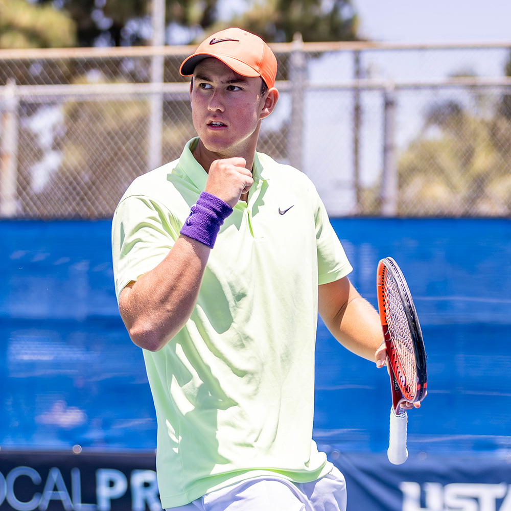 TCU Horned Frogs tennis player Oliver Bonding celebrates a point with a pumped fist during a match at the SoCal Pro Series in Lakewood. He is captured looking to the side with a determined expression, wearing a light neon-green Nike polo shirt, white shorts, an orange Nike cap, and a purple wristband on his right arm. He holds his tennis racket low in his left hand. In the background are blue court windscreens displaying banners for Wilson, SoCal Pro Series, and USTA Pro Circuit under bright, direct sunlight.
