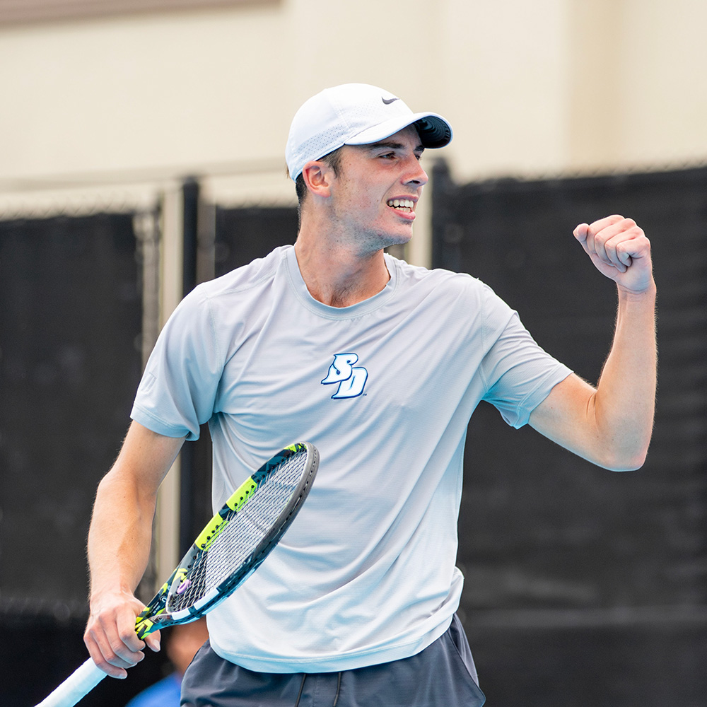University of San Diego tennis player Ollie Tarvet celebrates a point with a pumped left fist during a match at the 2025 SoCal Pro Series event hosted on his home court at the University of San Diego. He is captured looking to the side with an intense, determined expression and bared teeth. He is wearing a light grey Nike t-shirt featuring the blue USD athletic logo, dark grey shorts, and a white Nike baseball cap. He holds his tennis racket low in his right hand. The background consists of dark grey windscreens lining the outdoor tennis courts.