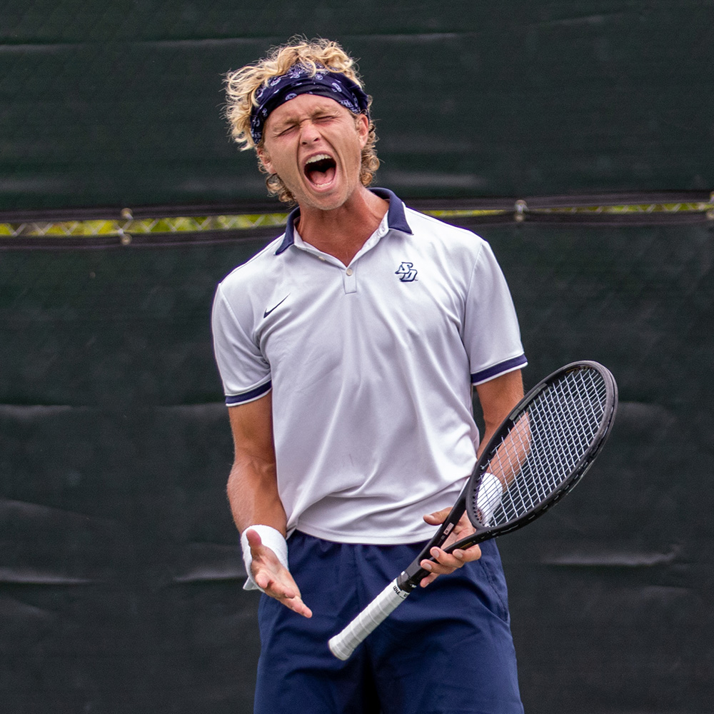 August Holmgren reacts with a powerful, passionate shout during his winning run at the 2022 SoCal Pro Series event hosted at the Rancho Santa Fe Tennis Club. He is captured with his eyes closed and mouth wide open in intense celebration, holding his tennis racket in his left hand. He is wearing a white collared Nike shirt featuring a USD (University of San Diego) logo, navy blue shorts, white wristbands, and a dark blue patterned bandana tied around his curly blonde hair. The background consists of a solid, dark green court windscreen.