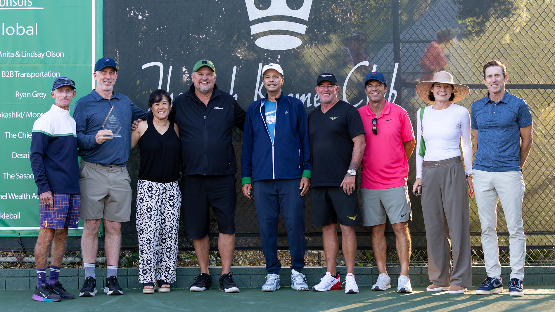 A group of nine key members of the Palos Verdes Peninsula tennis community pose together for a photo on an outdoor court at the Jack Kramer Club. Standing in order from left to right are Bob Krueger, Peter Smith (holding a small glass trophy), Kimberly Po, Trevor Kronemann, John Carrizosa, Oliver Messerli, Kip Brady, Aisling Bowyer, and Wade Murphy. They are lined up shoulder-to-shoulder, smiling in front of a dark court windscreen featuring the club's white crown logo. To the far left, a large green sign displays a list of tournament sponsors, including ITF Pro Circuit logos at the top.