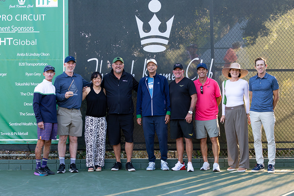 A group of nine key members of the Palos Verdes Peninsula tennis community pose together for a photo on an outdoor court at the Jack Kramer Club. Standing in order from left to right are Bob Krueger, Peter Smith (holding a small glass trophy), Kimberly Po, Trevor Kronemann, John Carrizosa, Oliver Messerli, Kip Brady, Aisling Bowyer, and Wade Murphy. They are lined up shoulder-to-shoulder, smiling in front of a dark court windscreen featuring the club's white crown logo. To the far left, a large green sign displays a list of tournament sponsors, including ITF Pro Circuit logos at the top.