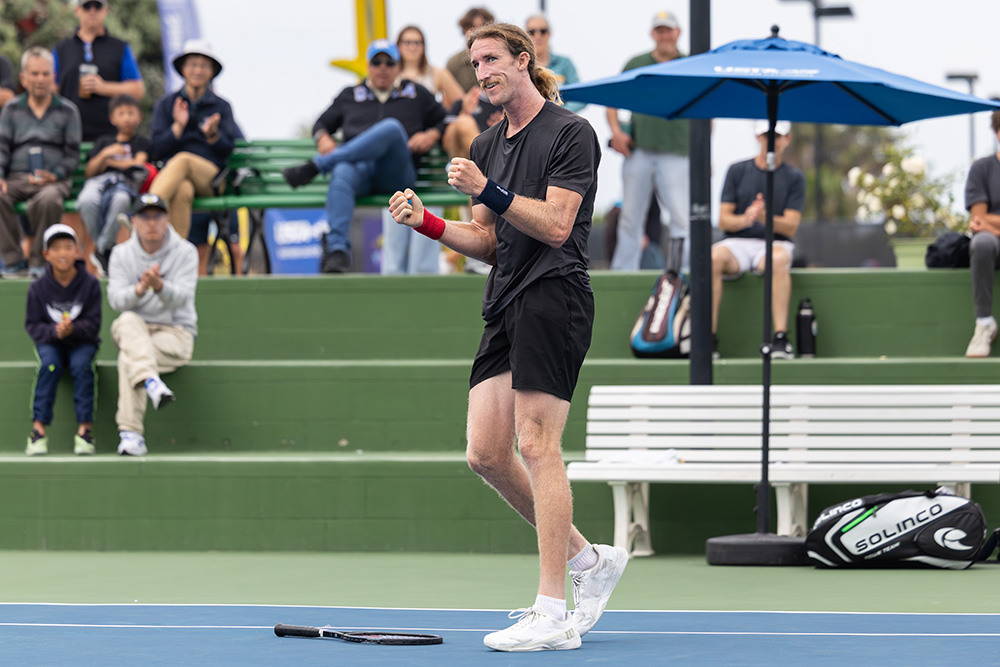 Keegan Smith celebrates winning the title at the 2025 SoCal Pro Series event held at the Barnes Tennis Center. He is captured smiling widely with both fists pumped in celebration after dropping his racket onto the blue hard court surface. He has a mustache and long blonde hair tied back, and he is wearing an all-black athletic kit consisting of a black t-shirt and black shorts, paired with white tennis shoes, a navy blue wristband on his left arm, and a red wristband on his right arm. In the blurred background, spectators clap from green stadium seating under a blue USTA umbrella.