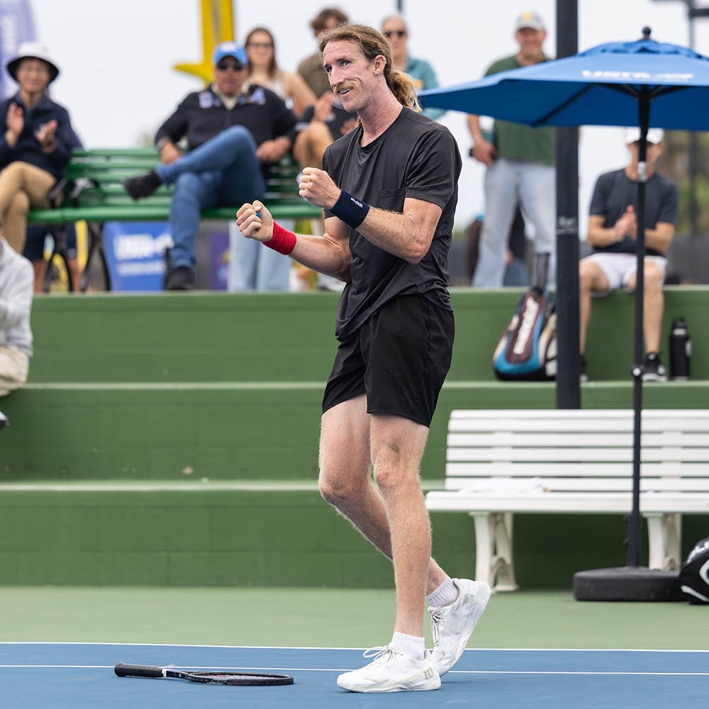 Keegan Smith celebrates winning the title at the 2025 SoCal Pro Series event held at the Barnes Tennis Center. He is captured smiling widely with both fists pumped in celebration after dropping his racket onto the blue hard court surface. He has a mustache and long blonde hair tied back, and he is wearing an all-black athletic kit consisting of a black t-shirt and black shorts, paired with white tennis shoes, a navy blue wristband on his left arm, and a red wristband on his right arm. In the blurred background, spectators clap from green stadium seating under a blue USTA umbrella.