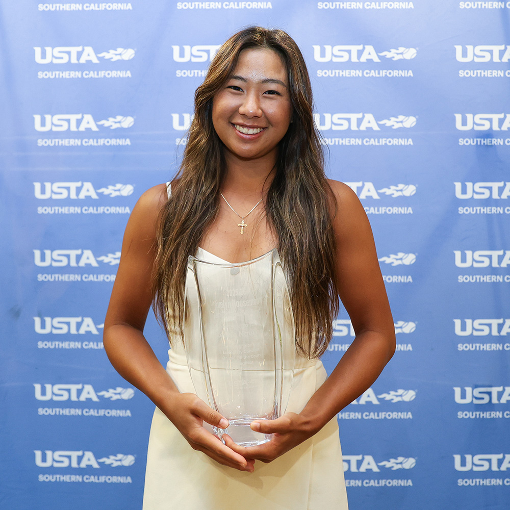 A portrait of a smiling Kenzie Nguyen holding a large, clear glass trophy with both hands after receiving the prestigious Evelyn Houseman Lifetime Sportsmanship Award. She has long, dark brown wavy hair, is wearing a white dress and a thin gold cross necklace, and stands in front of a blue backdrop patterned repeatedly with the USTA Southern California logo.
