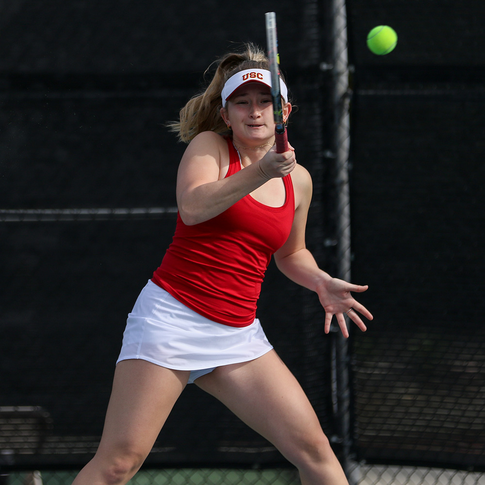 Tennis player Elizabeth Stuart-Chaffoo tracks a high-bouncing yellow ball while preparing to hit a forehand shot during the 2023 TOC SoCal Sectionals. She is captured in an athletic, focused stance on the court, wearing a red USC tank top, a white tennis skirt, and a white visor featuring red USC lettering. Her right arm is raised as she brings her racket forward, while her left hand is extended for balance. A black chain-link fence windscreens the background of the court.