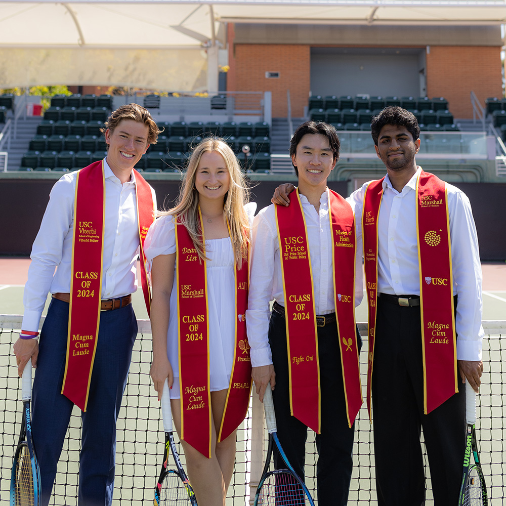 Four USC graduating seniors smile together for a portrait on a tennis court, standing shoulder-to-shoulder behind the net. The group consists of three men and one woman wearing white collared shirts or a white dress, paired with cardinal and gold USC graduation stoles indicating the Class of 2024. Each graduate is holding a tennis racket that rests against the court surface. The individual stoles display honors such as "Magna Cum Laude," "Dean's List," and specific schools including USC Viterbi, USC Marshall, and USC Price. In the background, the empty green stadium seating and structure of the tennis center are visible under bright daylight.