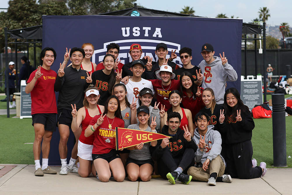 The USC Tennis On Campus team poses for a group photo at the 2023 TOC Sectionals at the Barnes Tennis Center. A large group of about twenty college athletes are gathered closely together, smiling and flashing "Fight On" victory signs with their fingers. They are dressed in various USC Trojans gear, including red, black, and grey t-shirts, sweatshirts, and tanks. Two players in the front center sit on the ground holding up a red and gold USC Trojans felt pennant flag. The group stands outdoors on a concrete pathway in front of a large navy blue USTA backdrop, with an open grassy area and palm trees visible in the background.