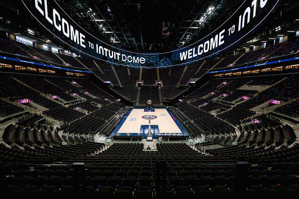 An interior, wide-angle view of the empty Intuit Dome basketball arena from the upper stands looking down at the court. The arena features thousands of dark seats rising in steep tiers around the polished wooden basketball court, which displays the LA Clippers logo at center court. High above the bowl, a massive, continuous double-sided halo display screen spans the perimeter of the ceiling, illuminated with the text "WELCOME TO INTUIT DOME".
