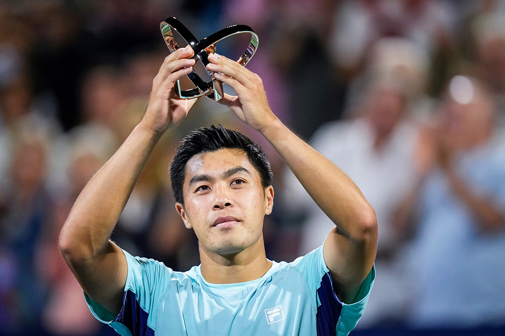A close-up portrait of tennis player Brandon Nakashima holding up his finalist trophy from the Montreal Open overhead with both hands. He is dressed in a light blue athletic shirt featuring the Fila logo and looks into the distance with a focused, proud expression against a softly blurred background.