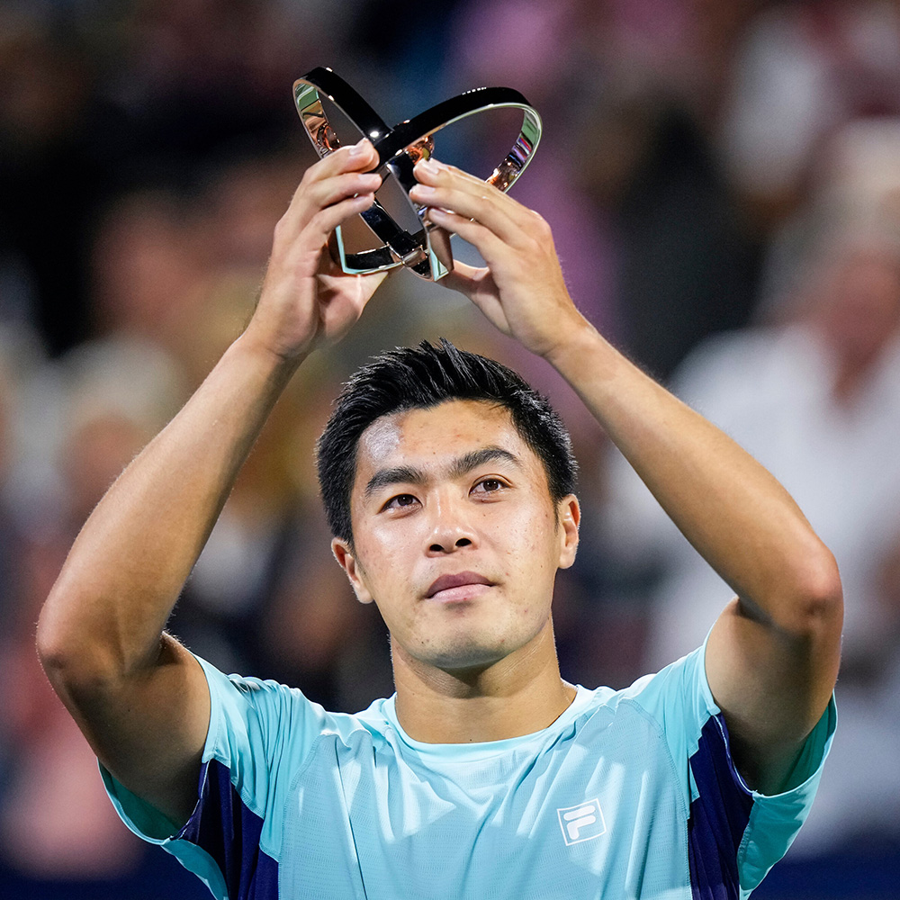 A close-up portrait of tennis player Brandon Nakashima holding up his finalist trophy from the Montreal Open overhead with both hands. He is dressed in a light blue athletic shirt featuring the Fila logo and looks into the distance with a focused, proud expression against a softly blurred background.