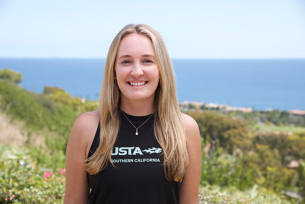 An outdoor headshot portrait of Riley Shear smiling directly at the camera in Palos Verdes. She has long, blonde hair and wears a black athletic tank top featuring the white USTA Southern California logo. A blurred view of lush green trees and the blue ocean horizon frames the background under a clear sky.