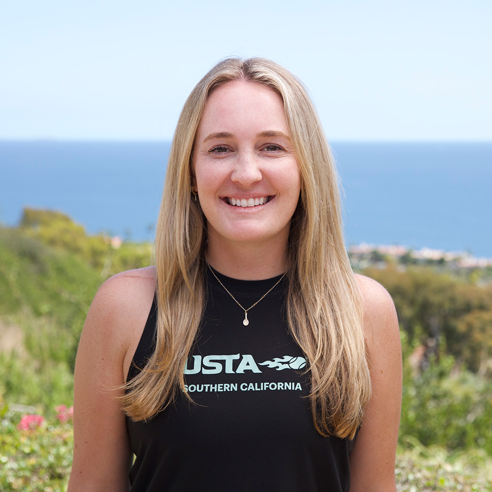 An outdoor headshot portrait of Riley Shear smiling directly at the camera in Palos Verdes. She has long, blonde hair and wears a black athletic tank top featuring the white USTA Southern California logo. A blurred view of lush green trees and the blue ocean horizon frames the background under a clear sky.
