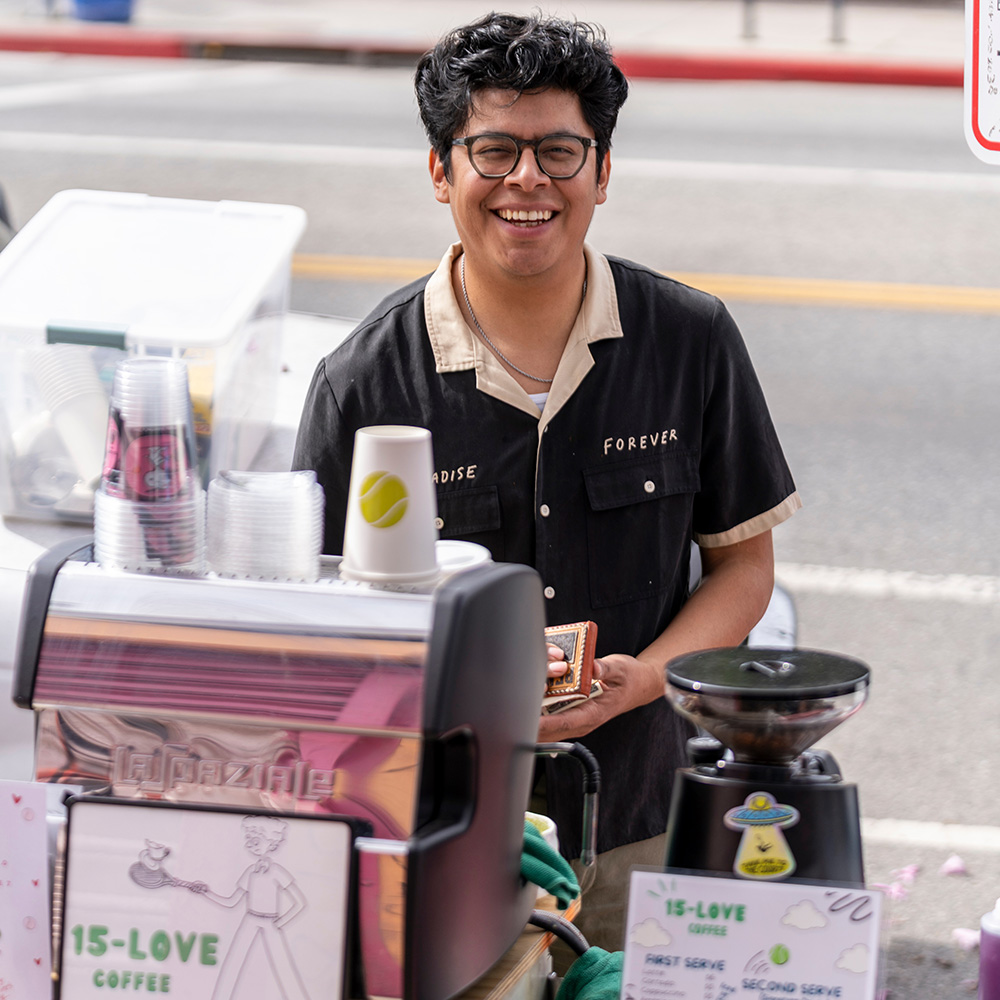 A smiling portrait of Bernie Parada standing behind a mobile coffee station for 15-LOVE Coffee. He wears glasses and a black button-up shirt with beige collar trim reading "PARADISE FOREVER." In the foreground, an espresso machine and coffee grinder are visible alongside branded cups featuring tennis ball art.