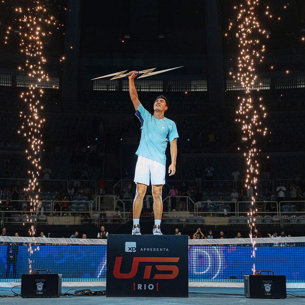 Tennis player Brandon Nakashima stands victorious atop a podium branded "UTS | RIO" after winning the Ultimate Tennis Showdown event. Dressed in a light blue Fila shirt and white shorts, he looks upward while holding a metallic lightning bolt trophy high in his right hand. Sparks shoot into the air from pyrotechnic fountains on either side of the podium inside the indoor arena.