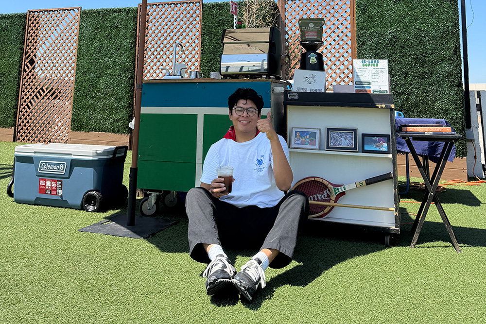 Bernie Parada sits cross-legged on artificial turf under a large white patio umbrella, smiling and giving a thumbs-up while holding an iced drink. He poses in front of his 15-LOVE Coffee cart setup, which features vintage tennis rackets, coffee equipment, a blue Coleman cooler, and a backdrop of green privacy hedging.