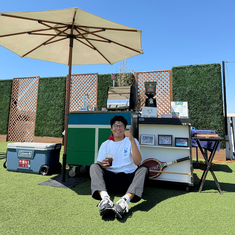 Bernie Parada sits cross-legged on artificial turf under a large white patio umbrella, smiling and giving a thumbs-up while holding an iced drink. He poses in front of his 15-LOVE Coffee cart setup, which features vintage tennis rackets, coffee equipment, a blue Coleman cooler, and a backdrop of green privacy hedging.