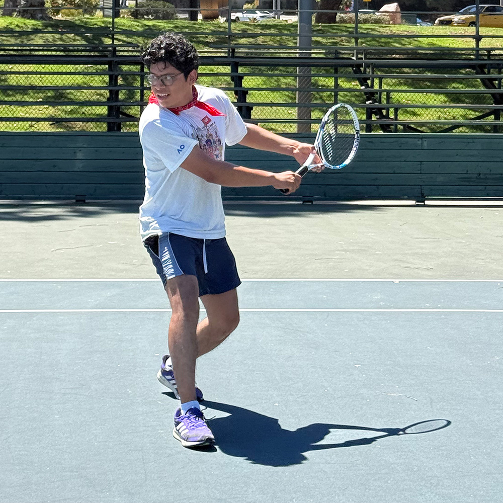 Bernie Parada in action on a hard tennis court, preparing to hit a two-handed backhand. He wears glasses, a white t-shirt, navy athletic shorts, purple sneakers, and a red bandana tied around his neck, with bleachers and greenery framing the outdoor court.