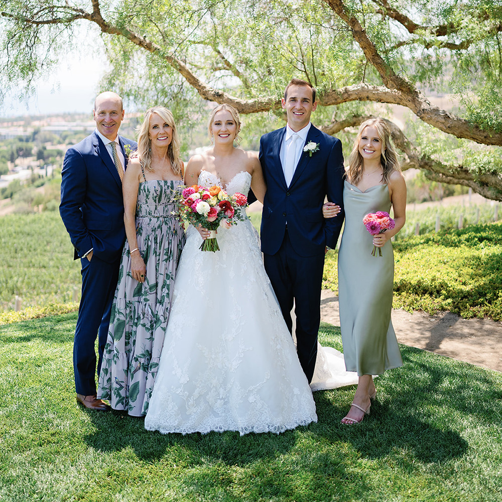 A outdoor wedding portrait featuring Riley Shear as the bride, standing in the center surrounded by her family under the shade of a large pepper tree. Riley wears a strapless white lace A-line gown and holds a colorful bouquet of pink, orange, and white flowers. Standing next to her is the groom in a navy blue suit, a bridesmaid in a sage green slip dress on the far right holding a small bouquet, and two parents dressed in formal wedding attire on the left. A scenic vineyard hillside stretches into the background under bright daylight.