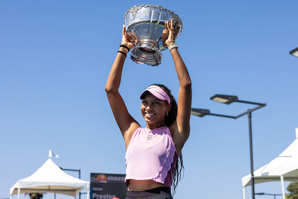 Tennis player Thea Frodin smiles brightly as she holds the large, ornate silver Maureen Connolly Brinker trophy high overhead with both hands after capturing the 18s singles title at the 2026 USTA Billie Jean King Girls' National Championships. She is captured from a low angle outdoors under a clear, bright blue sky, wearing a sleeveless light pink Nike crop top, a black tennis skirt, and a matching pink Nike visor over her long, dark curly hair. White event tents, outdoor court lighting fixtures, and spectating fans in red hats are visible at the base of the frame.