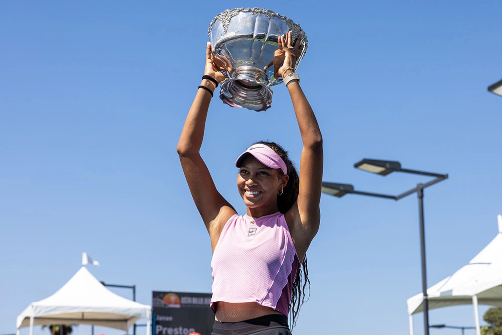 Tennis player Thea Frodin smiles brightly as she holds the large, ornate silver Maureen Connolly Brinker trophy high overhead with both hands after capturing the 18s singles title at the 2026 USTA Billie Jean King Girls' National Championships. She is captured from a low angle outdoors under a clear, bright blue sky, wearing a sleeveless light pink Nike crop top, a black tennis skirt, and a matching pink Nike visor over her long, dark curly hair. White event tents, outdoor court lighting fixtures, and spectating fans in red hats are visible at the base of the frame.