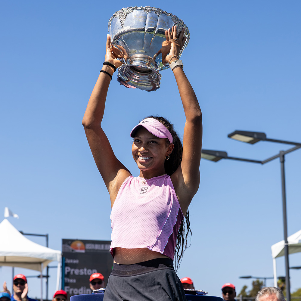 Tennis player Thea Frodin smiles brightly as she holds the large, ornate silver Maureen Connolly Brinker trophy high overhead with both hands after capturing the 18s singles title at the 2026 USTA Billie Jean King Girls' National Championships. She is captured from a low angle outdoors under a clear, bright blue sky, wearing a sleeveless light pink Nike crop top, a black tennis skirt, and a matching pink Nike visor over her long, dark curly hair. White event tents, outdoor court lighting fixtures, and spectating fans in red hats are visible at the base of the frame.