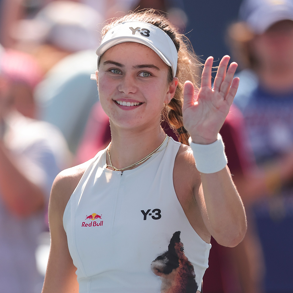 Tennis player Iva Jovic smiles and waves to the crowd following her victory at the 2025 US Open in Flushing, NY. She is captured in a close-up portrait wearing a white Y-3 sleeveless top with a Red Bull logo, a white Y-3 visor, a white wristband on her raised left hand, and a gold necklace, set against a softly blurred background of spectators and photographers.