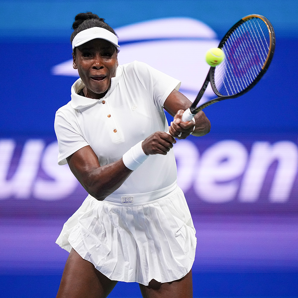 Venus Williams hits a two-handed backhand during a tennis match at the US Open. She wears an all-white athletic outfit, white visor, and wristbands against a purple court backdrop.