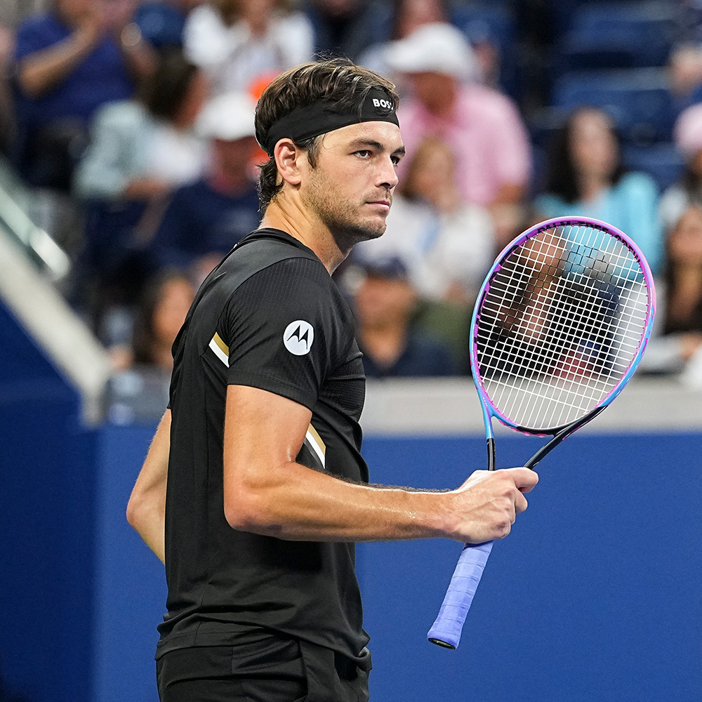 Taylor Fritz stands focused on the US Open court holding his tennis racket in profile view. He wears a black athletic shirt, a black BOSS headband, and holds a racket with a pink-and-blue frame.
