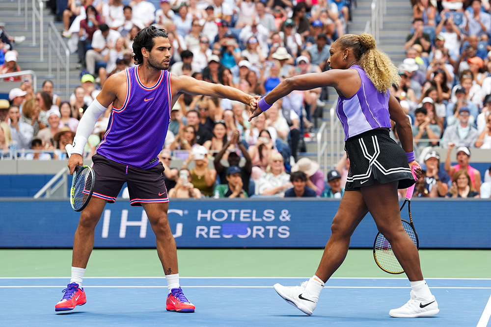 Mixed doubles partners Carlos Alcaraz and Serena Williams share a fist bump on a hard court during a tennis match. Carlos, on the left, wears a vibrant purple sleeveless Nike jersey with red and white trim, dark shorts, white compression sleeves, and bright red shoes while holding his tennis racket low in his right hand. Serena, on the right, reaches out to bump fists while wearing a sleeveless light-and-dark purple Nike top, a black pleated tennis skirt with white accent trim, and white Nike shoes, holding her racket behind her. They are surrounded by a packed stadium crowd filled with spectators under bright outdoor stadium lights.