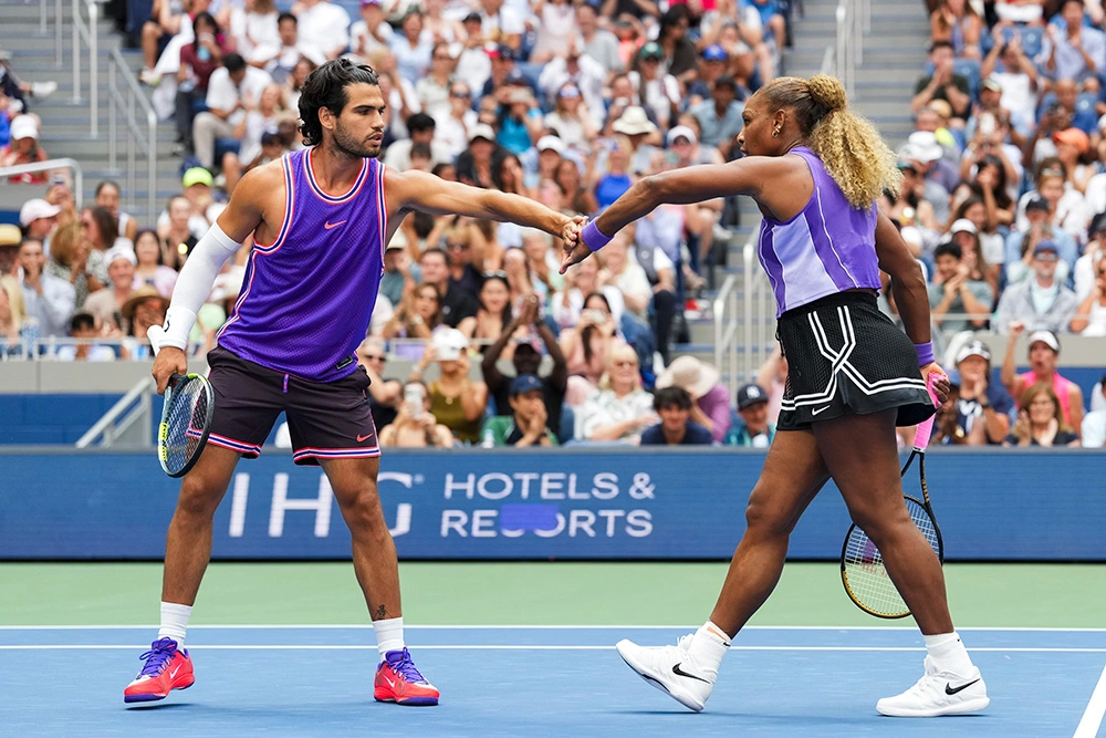 Mixed doubles partners Carlos Alcaraz and Serena Williams share a fist bump on a hard court during a tennis match. Carlos, on the left, wears a vibrant purple sleeveless Nike jersey with red and white trim, dark shorts, white compression sleeves, and bright red shoes while holding his tennis racket low in his right hand. Serena, on the right, reaches out to bump fists while wearing a sleeveless light-and-dark purple Nike top, a black pleated tennis skirt with white accent trim, and white Nike shoes, holding her racket behind her. They are surrounded by a packed stadium crowd filled with spectators under bright outdoor stadium lights.