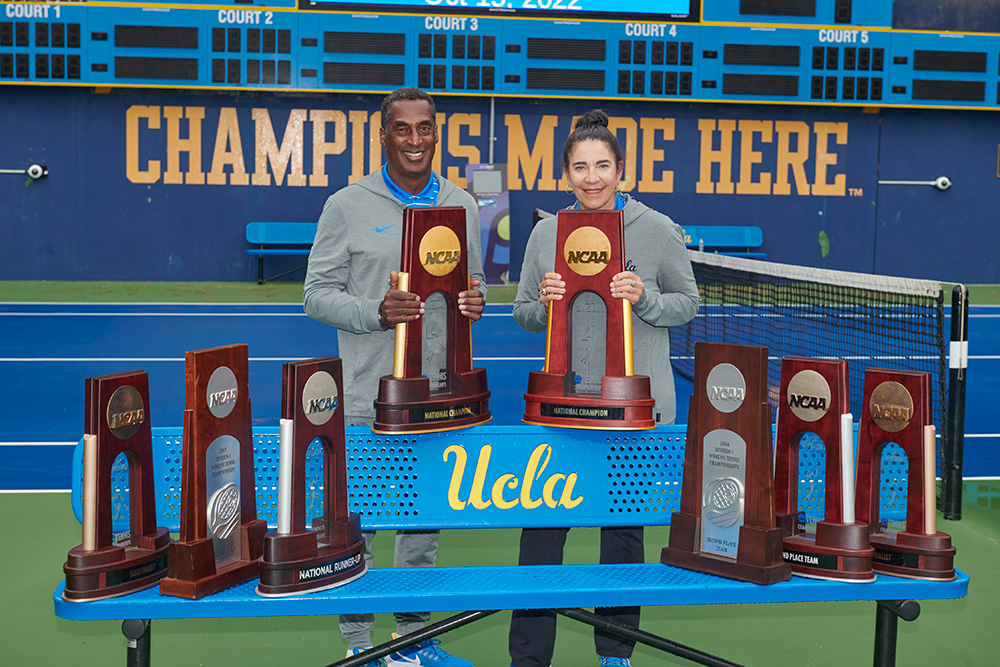 UCLA Women's Tennis head coach Stella Sampras Webster and associate head coach Rance Brown pose together behind a table displaying eight NCAA team and individual trophies. They stand outdoors on a blue hard court with a UCLA scoreboard reading "CHAMPIONS MADE HERE" in the background.