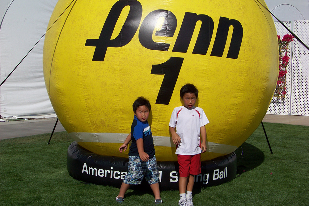 Young brothers Brandon and Bryce Nakashima pose together as children in front of a giant inflatable yellow tennis ball labeled "Penn 1". Bryce, on the left, wears a blue-and-black top with patterned shorts, while Brandon stands on the right in a white Nike shirt and red shorts on a grassy lawn.