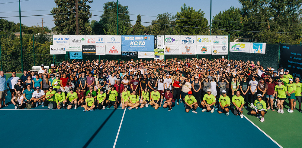 A large group of dozens of tennis players, coaches, and community members pose together on a bright blue outdoor tennis court during a Kern Community Tennis Association (KCTA) event. A long backdrop fence behind them is covered with sponsor banners, including USTA, Head, and KCTA logos.