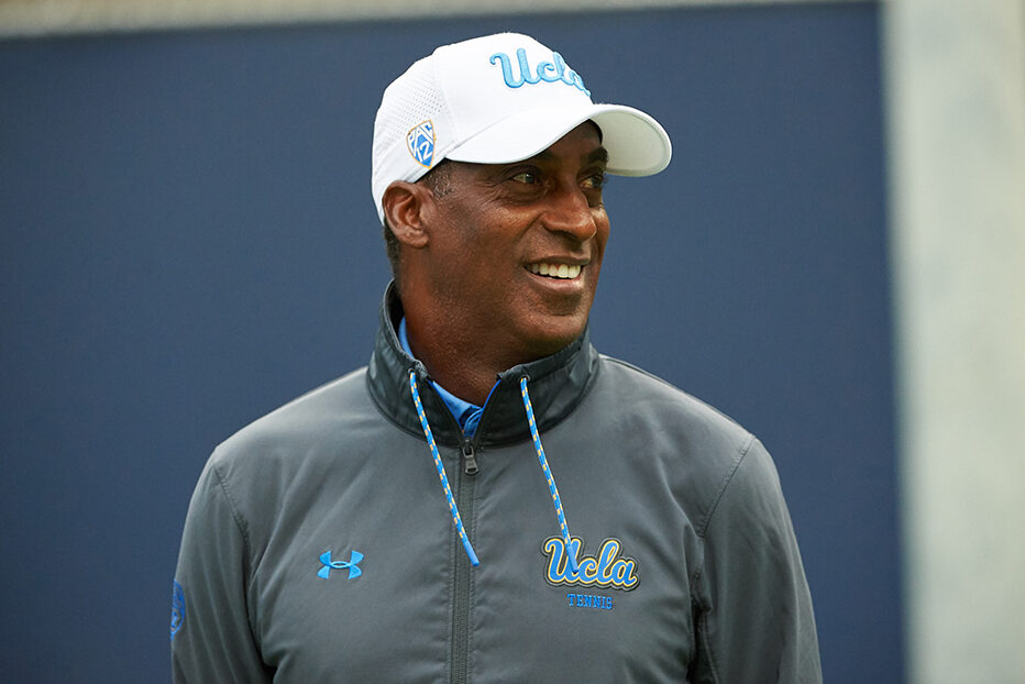 A outdoor portrait of UCLA associate head coach Rance Brown smiling in profile. He wears a grey quarter-zip UCLA Tennis jacket and a white UCLA cap featuring the Pac-12 logo on the side.