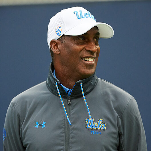 A outdoor portrait of UCLA associate head coach Rance Brown smiling in profile. He wears a grey quarter-zip UCLA Tennis jacket and a white UCLA cap featuring the Pac-12 logo on the side.
