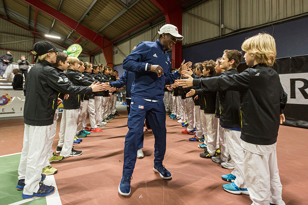 Rance Brown runs down a high-five line formed by young junior tennis players inside an indoor court facility. Dressed in a navy USA Tennis tracksuit and white cap, he smiles as he exchanges high-fives with kids standing in two parallel rows.
