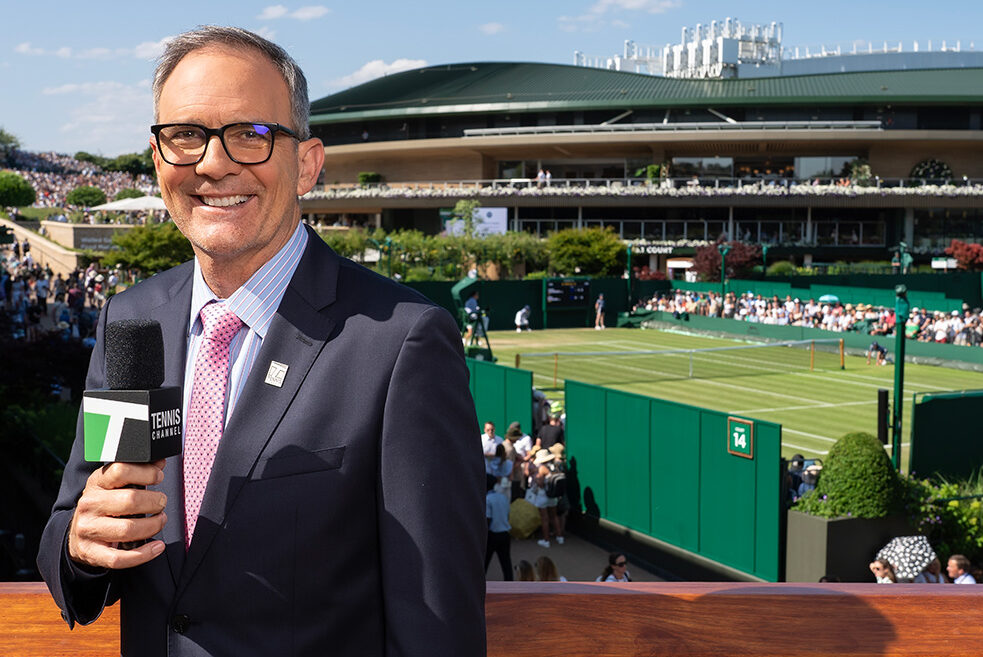 Tennis commentator and former professional player Paul Annacone smiles holding a Tennis Channel microphone on set during live broadcast coverage. He wears a navy suit jacket, a light blue striped button-down shirt, a patterned pink tie, and black-rimmed glasses. Behind him, the lush green grass courts and iconic stadium architecture of Wimbledon are visible under a bright, clear sky.