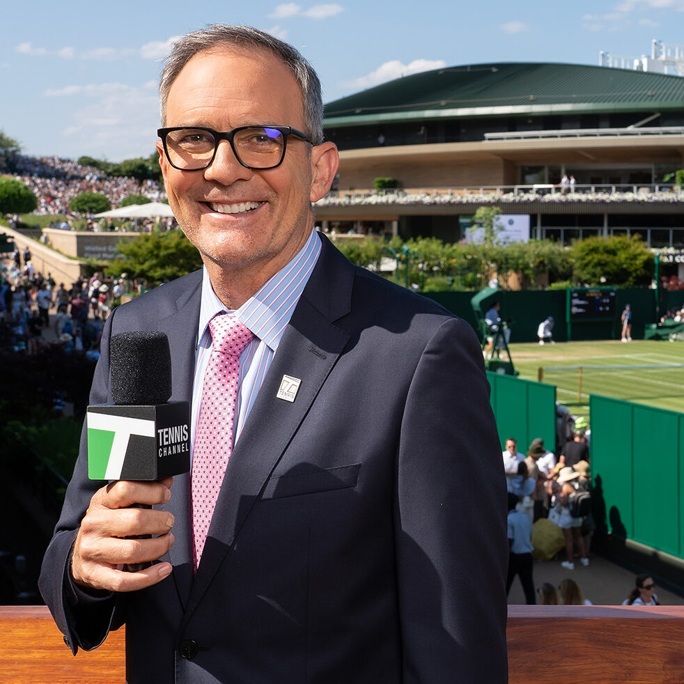 Tennis commentator and former professional player Paul Annacone smiles holding a Tennis Channel microphone on set during live broadcast coverage. He wears a navy suit jacket, a light blue striped button-down shirt, a patterned pink tie, and black-rimmed glasses. Behind him, the lush green grass courts and iconic stadium architecture of Wimbledon are visible under a bright, clear sky.