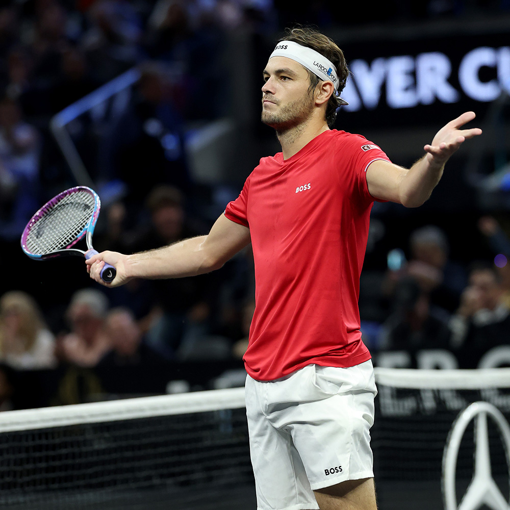 American tennis player Taylor Fritz gestures in celebratory disbelief after defeating Carlos Alcaraz at the 2025 Laver Cup in San Francisco. Dressed in Team World's red Boss t-shirt, white shorts, and a white headband, he holds his tennis racket in his right hand while holding out his left palm toward the crowd with an intense, triumphant expression. The iconic black Laver Cup court surround and blurred spectators fill the background behind the net.