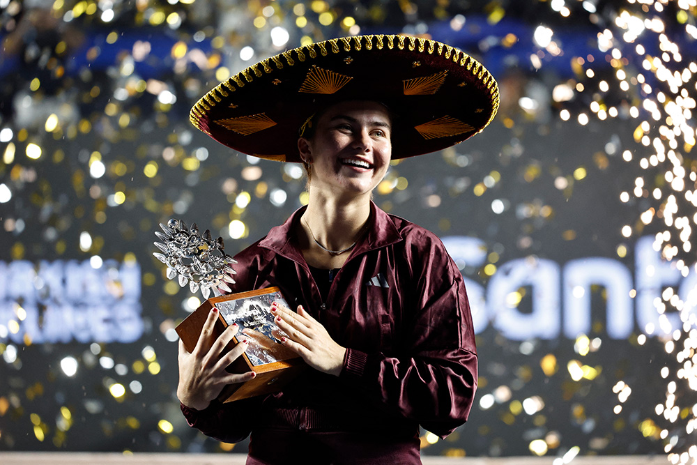 Tennis player Iva Jovic smiles happily as she holds her silver pineapple-topped Guadalajara Open championship trophy while wearing a traditional black and gold embroidered Mexican sombrero. She is dressed in a dark maroon Adidas jacket, standing in celebration as gold confetti falls around her against a dark, bokeh-lit background during the trophy ceremony.