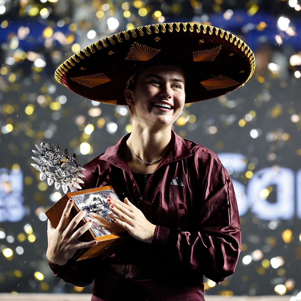 Tennis player Iva Jovic smiles happily as she holds her silver pineapple-topped Guadalajara Open championship trophy while wearing a traditional black and gold embroidered Mexican sombrero. She is dressed in a dark maroon Adidas jacket, standing in celebration as gold confetti falls around her against a dark, bokeh-lit background during the trophy ceremony.