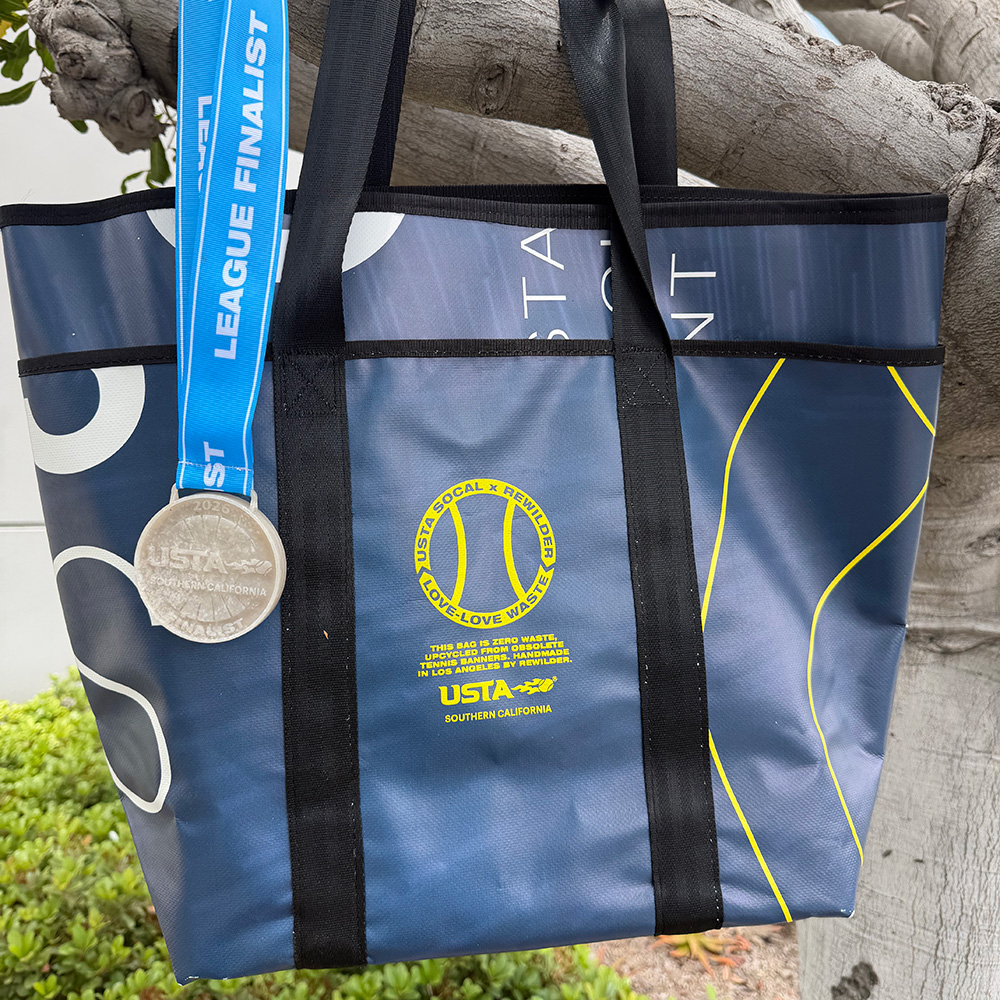 A durable blue tote bag crafted by Rewilder from upcycled, obsolete USTA Southern California tennis banners. The bag features black seatbelt-style carrying straps and a printed circular emblem that reads "USTA SOCAL x REWILDER | LOVE-LOVE WASTE," along with text noting its zero-waste design. Also an Adult League finalist medal.