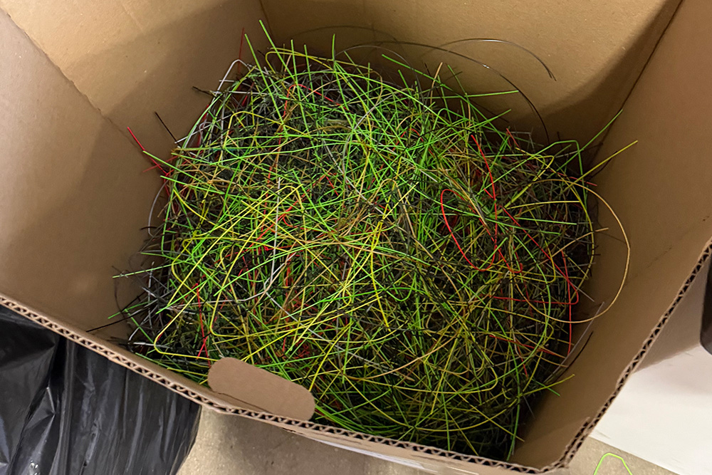 A top-down view into a cardboard collection box filled with a dense bundle of used, colorful tennis racket strings—primarily neon green, red, black, and white—gathered to be repurposed for LET.Tennis.
