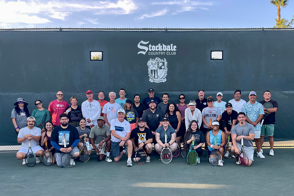 A group photo of tennis players posing on an outdoor court at the Stockdale Country Club. Jerry Matthews stands in the front row, sixth from the left, wearing a white t-shirt and hat while smiling alongside fellow players lined up against a dark green court screen displaying the club's crest and name.