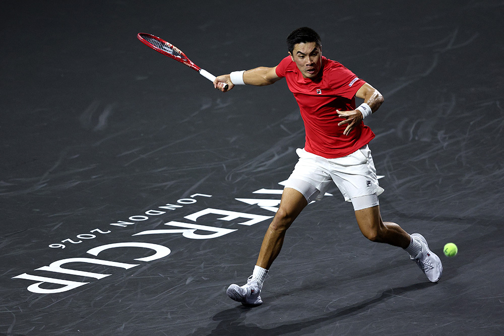 Brandon Nakashima of Team World stretches out wide across the court to play a forehand against Jakub Mensik of Team Europe during the Day 1 Men's Singles match at the 2026 Laver Cup at The O2 in London. He wears a red Fila shirt, white shorts, white wristbands, and white shoes, captured mid-stride on the signature dark grey hard court painted with the white "LAVER CUP LONDON 2026" tournament logo.